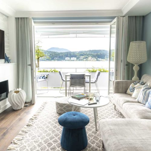 Wohnzimmer mit gemütlichem Sofa, Blick auf einen Balkon und See im Hintergrund, dekoriert mit blauen Akzenten.