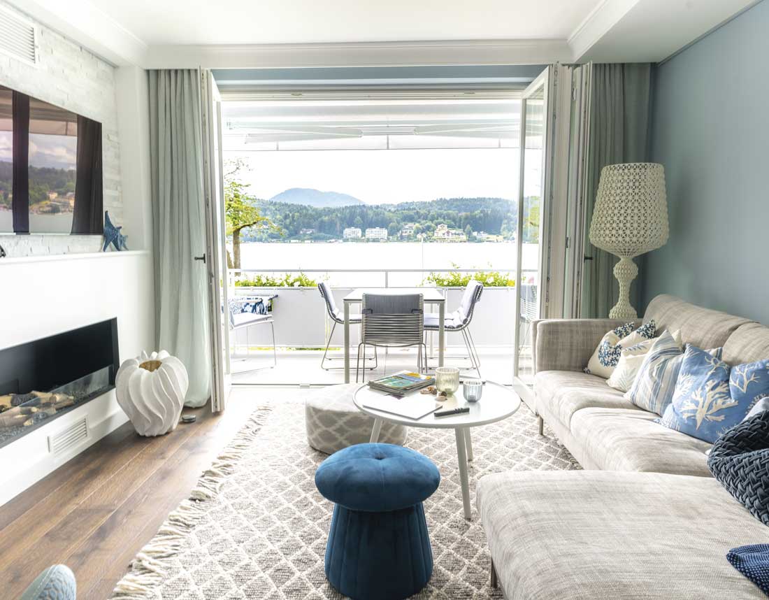 Wohnzimmer mit gemütlichem Sofa, Blick auf einen Balkon und See im Hintergrund, dekoriert mit blauen Akzenten.